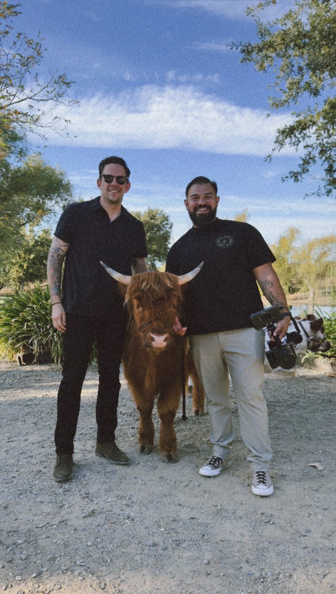 Two filmmakers outdoors with a highland cow, one holding a camera rig